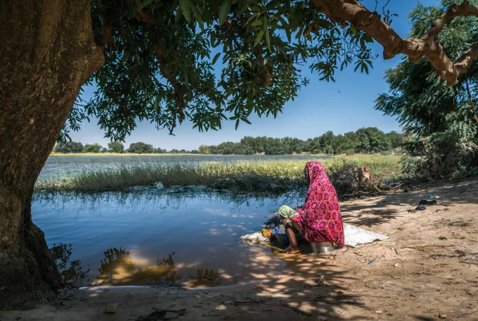 A woman is sat at the edge of a lake whilst washing clothes